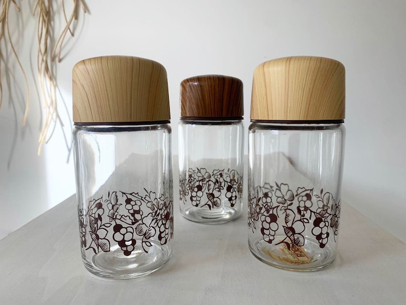 May include: Three glass jars with wooden lids and a grapevine design. The jars are clear glass with a brown grapevine design around the middle. The lids are a light wood color.