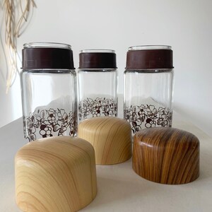 May include: Three glass jars with brown lids and a floral design. Each jar has a wooden lid with a woodgrain pattern. The lids are in two colors: light brown and dark brown.