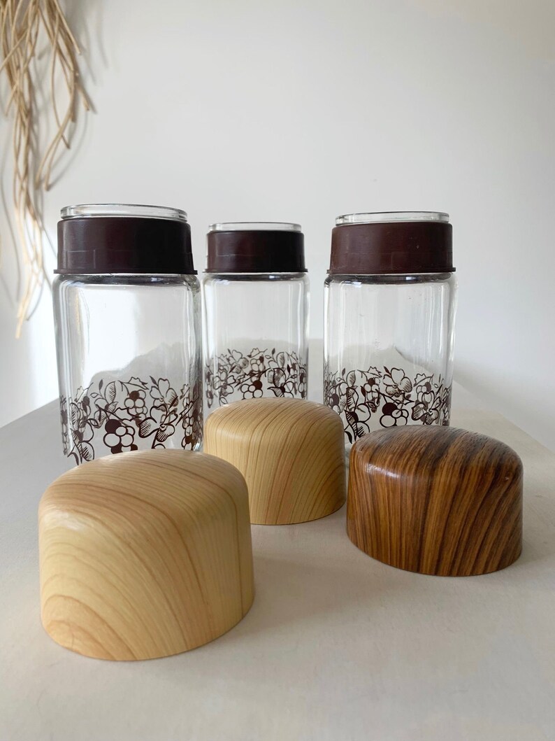 May include: Three glass jars with brown lids and a floral design. Each jar has a wooden lid with a woodgrain pattern. The lids are in two colors: light brown and dark brown.