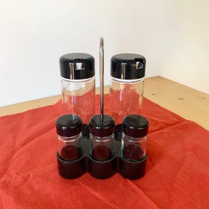 May include: A black metal spice rack with five glass bottles. The bottles have black lids and are filled with spices. The rack is sitting on a red cloth.