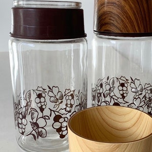 May include: Two glass jars with brown lids and a grapevine design. The jars have a wooden lid on top. A wooden bowl is in the foreground.
