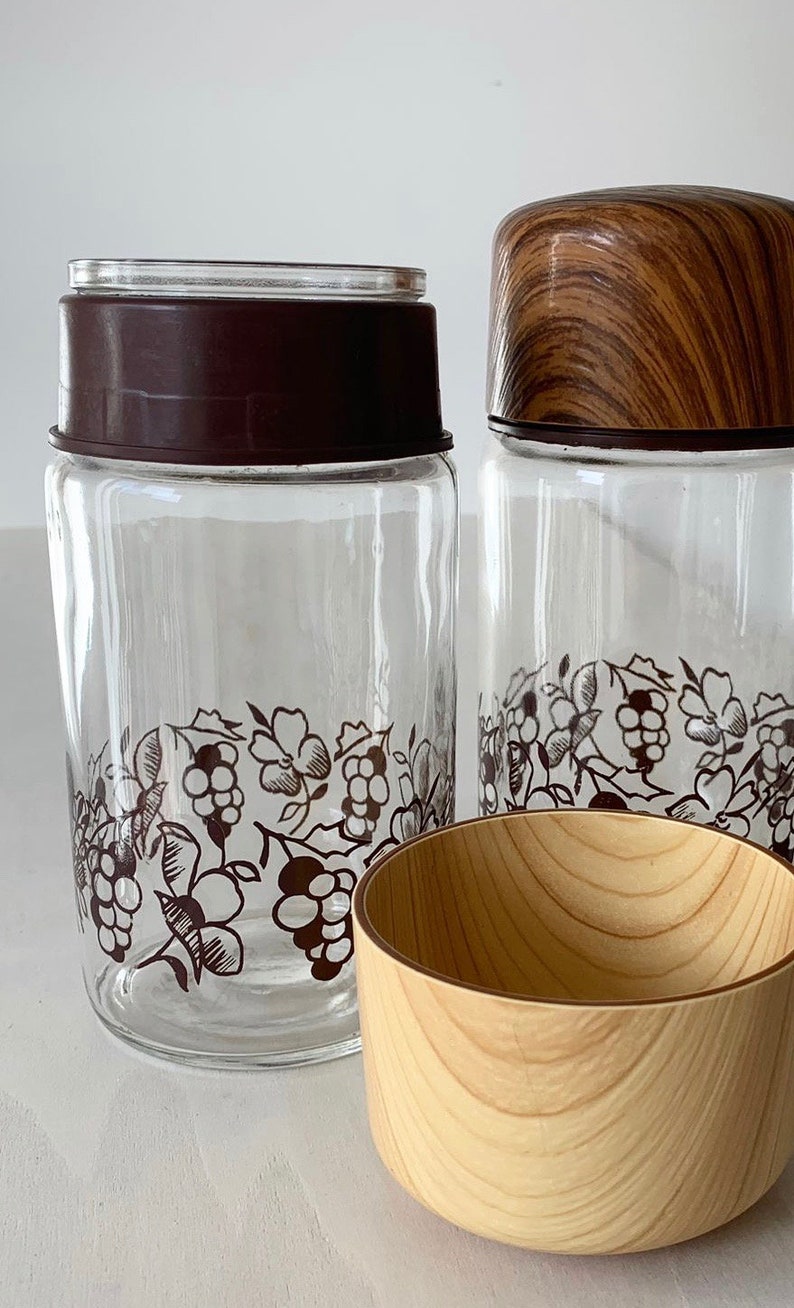 May include: Two glass jars with brown lids and a grapevine design. The jars have a wooden lid on top. A wooden bowl is in the foreground.