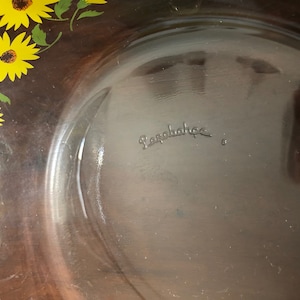 May include: A clear glass baking dish with a brown rim and a sunflower design. The bottom of the dish is marked with the word "Pyrex".