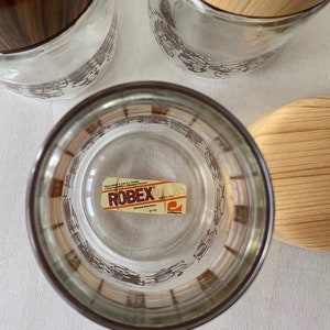 May include: Set of three glass storage jars with brown and white floral designs. The jars have wooden lids with a brown wood grain pattern. The jars have a label that reads "ROBEK" with the text "Chiusure Bresciane" and "Made in Italy".