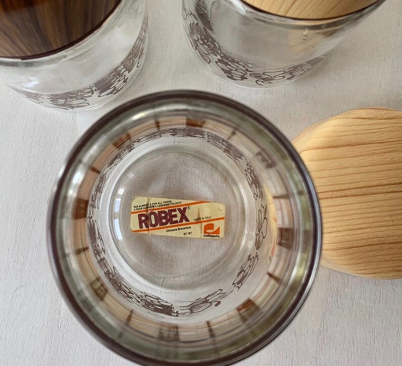 May include: Set of three glass storage jars with brown and white floral designs. The jars have wooden lids with a brown wood grain pattern. The jars have a label that reads "ROBEK" with the text "Chiusure Bresciane" and "Made in Italy".