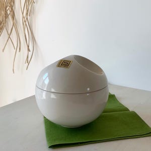 Op de afbeelding: Witte keramische kom met een deksel, rond van vorm en met een kleine opening aan de bovenkant. De kom heeft een gouden label met de tekst "SOLE-ARTE".