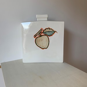 Könnte beinhalten: Weiße Keramikvase mit einem stilisierten Apfeldesign in Orange, Grün und Braun. Der Apfel hat ein Blatt und einen Stiel.