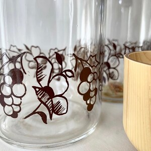 May include: Four clear glass tumblers with a black and white grapevine design around the top. The glasses are sitting on a white surface next to a wooden bowl.