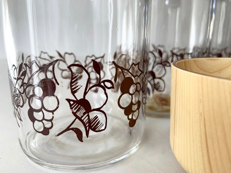 May include: Four clear glass tumblers with a black and white grapevine design around the top. The glasses are sitting on a white surface next to a wooden bowl.