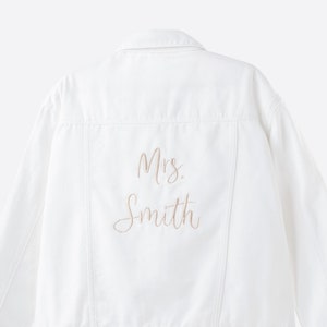 May include: A white denim jacket with the embroidered text "Mrs. Smith" in a cursive font. The jacket has silver buttons and a classic design, perfect for a wedding or bridal event.