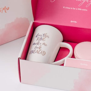 May include: A pink gift box with a white mug that says "Does this ring make me look engaged?" in purple and gold lettering. The mug is in a pink box with a white lid. The box is decorated with a watercolor-style pink and white design.