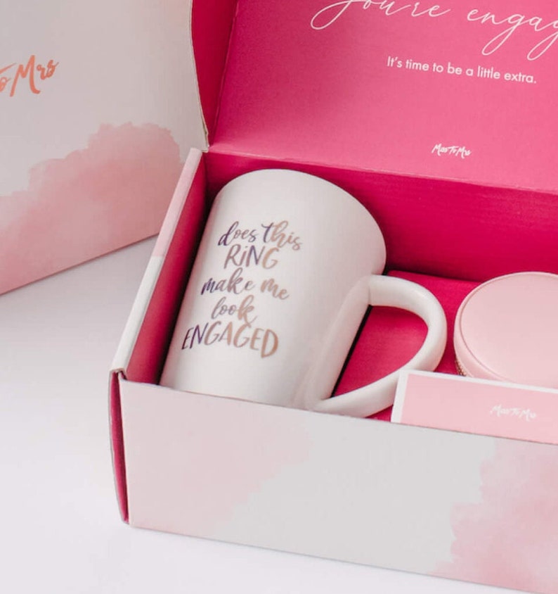 May include: A pink gift box with a white mug that says "Does this ring make me look engaged?" in purple and gold lettering. The mug is in a pink box with a white lid. The box is decorated with a watercolor-style pink and white design.