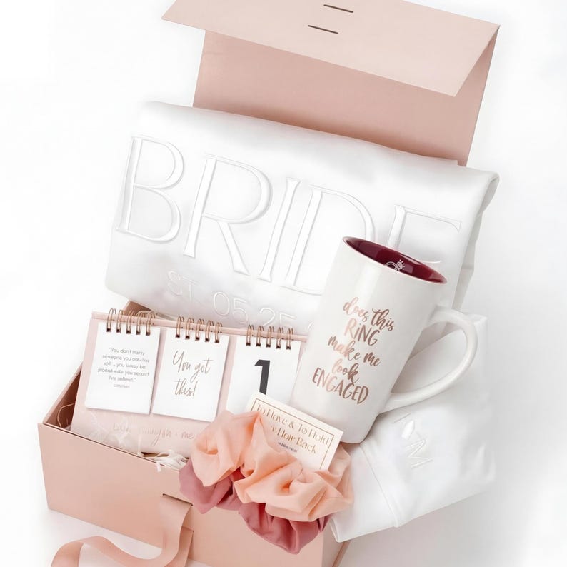 以下が含まれることがあります： 「BRIDE」のエンボス加工が施された白い枕、"does this RING make me look ENGAGED"と書かれたマグカップ、カレンダー、シュシュ、カードが入ったピンク色のギフトボックス。ボックスは開いており、中身が見えています。
