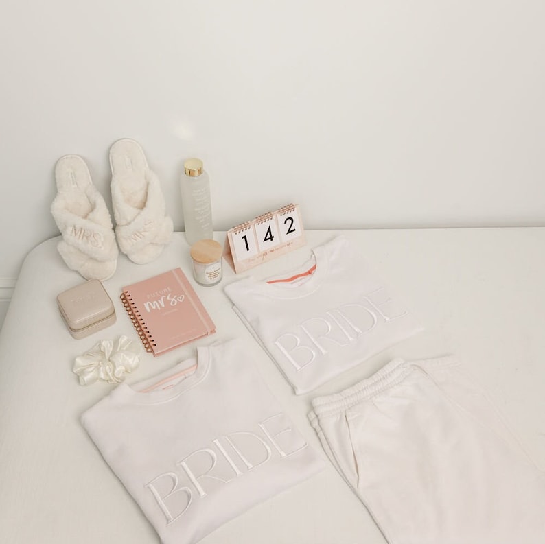 May include: White bridal pajamas with the word "Bride" embroidered on the shirts and pants. The set includes a pair of white slippers with the word "Mrs" embroidered on them, a pink notebook with the words "Future Mrs" on the cover, a small white candle, a clear glass bottle, and a small calendar with the numbers "142" displayed.