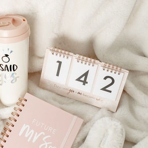 May include: A pink countdown calendar with the numbers 142 displayed. The calendar reads "Days until you + me become we". A pink notebook with the words "Future Mrs" is in the foreground. A pink and white travel mug with the words "I said yes" is in the background.