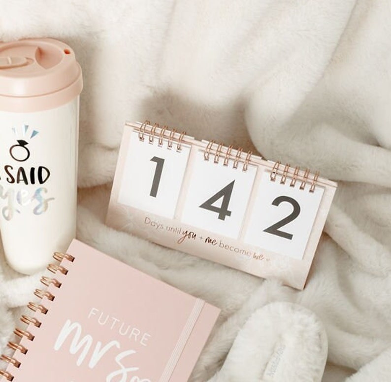 May include: A pink countdown calendar with the numbers 142 displayed. The calendar reads "Days until you + me become we". A pink notebook with the words "Future Mrs" is in the foreground. A pink and white travel mug with the words "I said yes" is in the background.