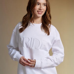 以下が含まれることがあります： 胸に「BRIDE」の文字が刺繍された白いスウェットシャツ。長袖トップスはクルーネックとリブ編みの袖口が付いています。袖口には小さな刺繍のディテールがあります。スウェットシャツは柔らかい素材で作られています。