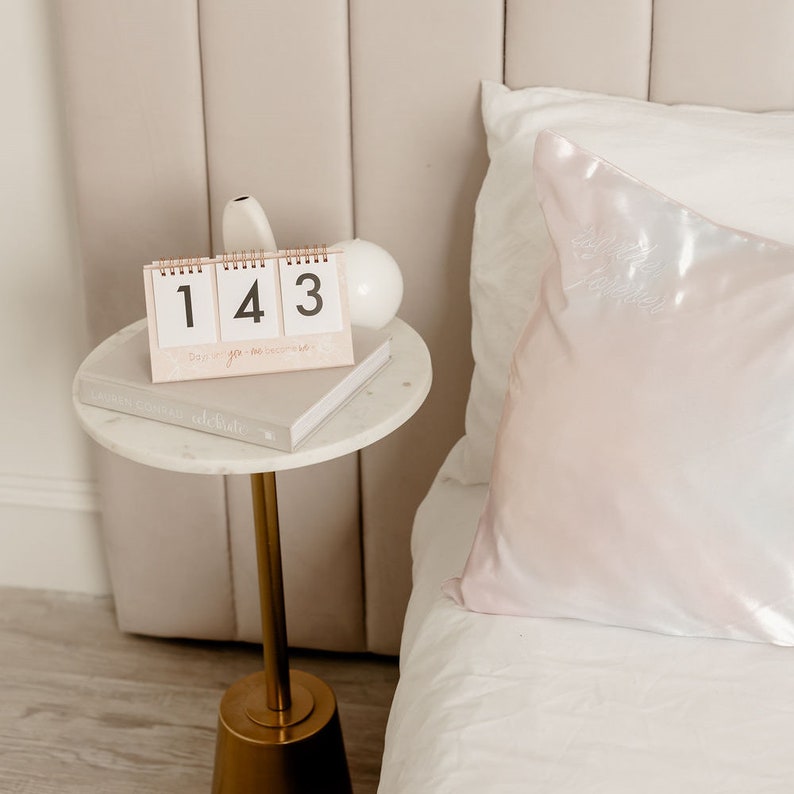 May include: A white bedside table with a gold metal stand and a marble top. A calendar with the numbers 143 is on the table, along with a book titled "Lauren Conrad Celebrate".