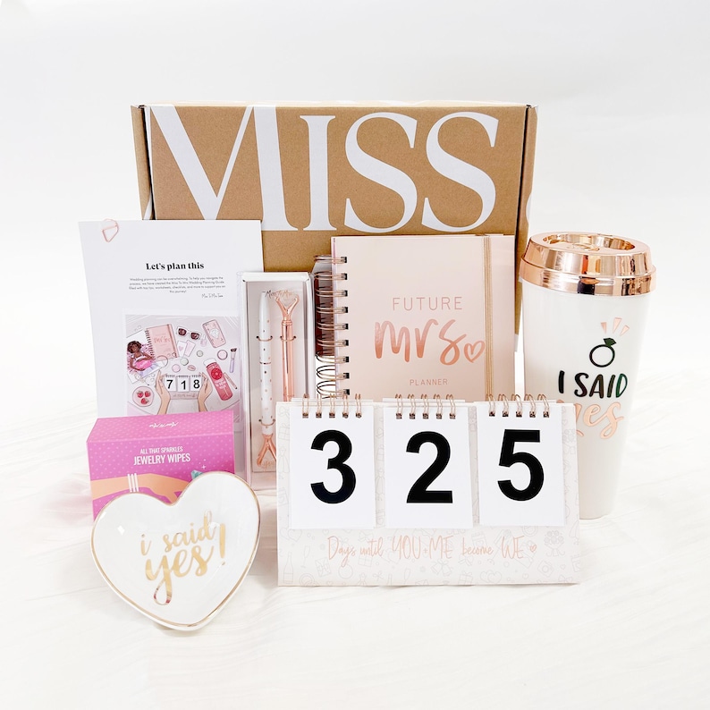 May include: A brown box with the word "MISS" printed on the front. The box contains a variety of wedding-themed items, including a planner, a countdown calendar, a mug, a jewelry dish, and a pen. The planner is pink and has the words "FUTURE MRS" on the front. The countdown calendar is white and has the numbers "325" on it. The mug is white with a gold rim and has the words "I SAID YES" on it. The jewelry dish is white and heart-shaped with the words "I SAID YES" on it. The pen is gold and has a diamond-shaped top.