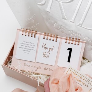 以下が含まれることがあります： ピンク色のギフトボックスには、「Days until you + me bec」のテキストが入ったカウントダウンカレンダーが入っています。 カレンダーには「You got this!」という言葉と数字「1」が書かれた3ページがあります。 また、シュシュと装飾枕も含まれています。