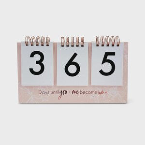 以下が含まれることがあります： 3つの白いカードに数字365が表示された、ピンクと白のカウントダウンカレンダー。カレンダーには花の模様があり、「Days until you + me become we」というテキストがあります。銅色のワイヤーでまとめられています。