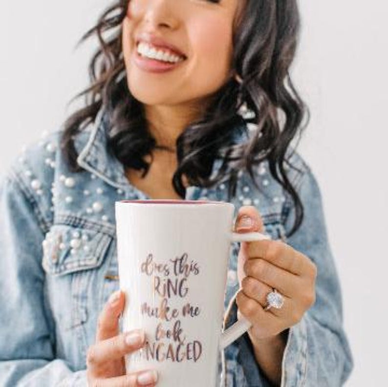 May include: A white ceramic coffee mug with a black and gold design that says "Does this ring make me look engaged?" A person is holding the mug and showing off a large diamond engagement ring on their finger.