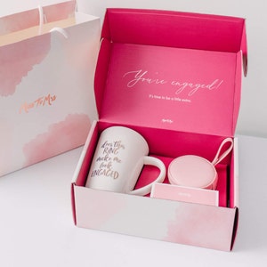 May include: A pink gift box with a white mug that says "Does this ring make me look engaged?" and a small pink ring box. The box is open and the lid has the text "You're engaged! It's time to be a little extra."