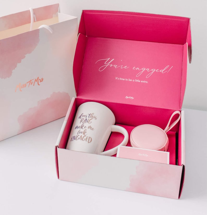 May include: A pink gift box with a white mug that says "Does this ring make me look engaged?" and a small pink ring box. The box is open and the lid has the text "You're engaged! It's time to be a little extra."