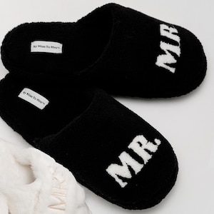 May include: A pair of black fuzzy slippers with white "MR." embroidery. The slippers are made by Miss To Mrs.