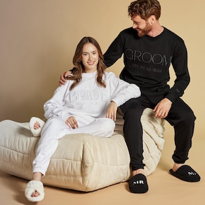 May include: A couple wearing matching white and black sweatshirts with the words "Bride" and "Groom" embroidered on them. The woman is sitting on a cream-colored beanbag chair and the man is standing behind her with his arm around her shoulder. They are both wearing matching black and white slippers with the words "Bride" and "Groom" embroidered on them.