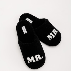 May include: A pair of black fuzzy slippers with the word "MR." embroidered in white on each slipper.