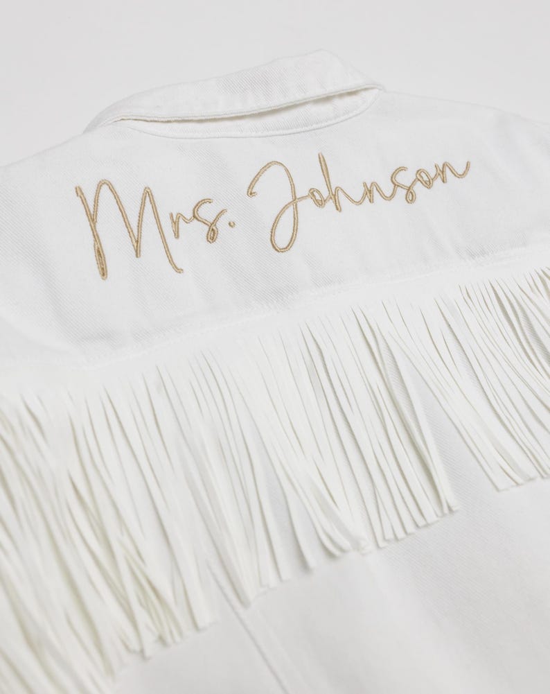 Może przedstawiać: Biała kurtka dżinsowa z frędzlami i haftowanym złotym napisem "Mrs. Johnson". Kurtka ma klasyczny kołnierz i zapięcie na guziki, z frędzlami z tyłu.