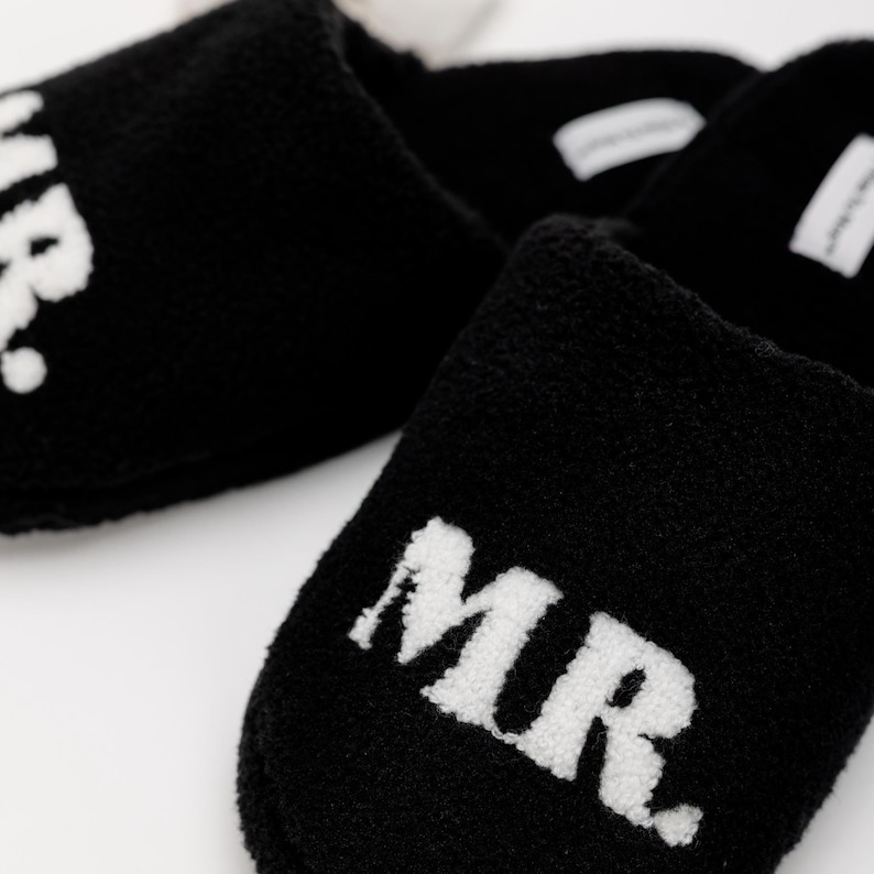 May include: A pair of black fuzzy slippers with white embroidered text. One slipper says "MR." and the other says "MRS."