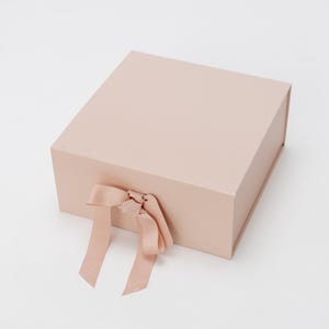 以下が含まれることがあります： ピンク色のギフトボックス。サテンのリボンが蝶々結びで結ばれています。ボックスは長方形で、滑らかでマットな仕上がりです。リボンが蝶々結びから垂れ下がっています。