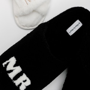 May include: Black fuzzy slippers with the word "MR" embroidered in white. The slippers have a white tag that says "by MISS TOMS."