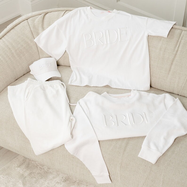 May include: White bride-to-be pajama set with a white t-shirt, sweatshirt, and baseball cap. Each piece features the word "BRIDE" in raised lettering.