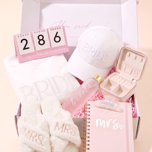 May include: A gift box containing a white sweatshirt with BRIDE text, a white baseball cap with BRIDE embroidery, a pink countdown calendar, a frosted water bottle with bride text, a small jewelry case, fuzzy white slippers with MRS text, and a notebook.