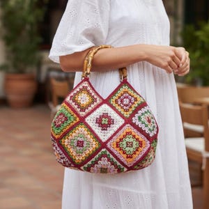 Op de afbeelding: Een handtas gemaakt van veelkleurige gehaakte granny squares in tinten bordeaux, groen, geel en wit. De tas heeft twee gebogen bamboe handvatten aan de bovenkant, wat zorgt voor een gestructureerde vorm met een levendig en ingewikkeld patroon.