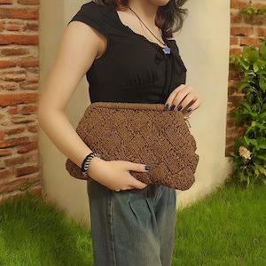 May include: A brown woven clutch bag with a zip fastening. The bag is being held by a person wearing a black top and blue jeans.