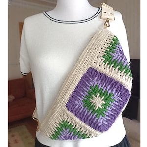 May include: A crossbody bag crafted from cream, purple, and green yarn in a square crochet pattern. The bag features a wide, adjustable cream-colored strap with gold-toned metal hardware, worn across the chest over a white short-sleeved shirt.