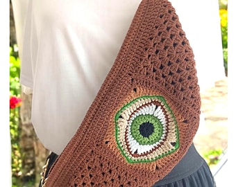 May include: A crescent-shaped brown crochet crossbody bag featuring a central eye-like motif with concentric rings of black, green, white, and tan yarn. The bag has a textured stitch pattern and is attached to a gold-toned metal chain strap.