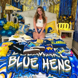 May include: A bed covered in University of Delaware Blue Hens merchandise. Items include a blanket, shirts, hats, shoes, and balloons in blue and yellow. The word "DELAWARE" is spelled out in silver balloons.