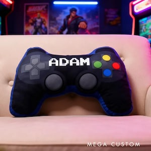 May include: A black and blue video game controller-shaped pillow with the name "ADAM" in white pixelated letters. The pillow has colorful buttons and is on a beige couch. The text "MEGA CUSTOM" is at the bottom.