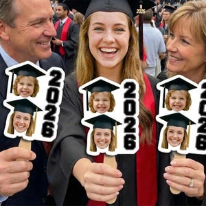 May include: People at a graduation ceremony holding custom photo props. Each prop features a graduation cap and the year "2026" in bold black letters. The props display two different photos of a young girl and a young woman.