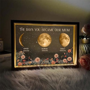May include: Illuminated framed art piece with the text "THE DAYS YOU BECAME OUR MUM." The artwork features three moon phases, names, and dates: David 08/20/1987, Sophia 10/05/1990, and Emily 02/05/1993. Floral accents adorn the bottom.