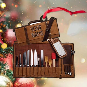 May include: A brown canvas chef's knife roll with a matching carrying case. The case has the text "YOUR NAME" embossed on it. The roll contains various knives, utensils, and a red spatula. A red ribbon is attached for hanging.