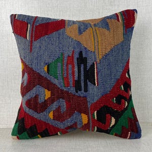 Può includere: Un cuscino quadrato con una fodera in tessuto intrecciato di colore blu di base. Il design presenta motivi geometrici in rosso, beige, verde e nero, con forme stilizzate e linee angolari distribuite sulla superficie anteriore.