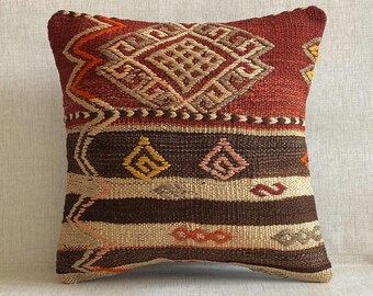 May include: A square throw pillow featuring a woven textile design with a deep red upper section containing a large diamond pattern. The lower portion consists of horizontal stripes in dark brown and beige, accented with small geometric motifs.
