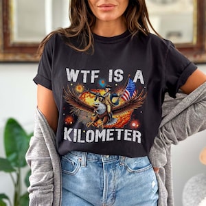 Può includere: Una maglietta nera con una grafica di un'aquila calva che trasporta un uomo vestito da soldato della guerra d'indipendenza americana. L'aquila tiene una bandiera americana e il testo "WTF IS A KILOMETER" è stampato sotto la grafica.
