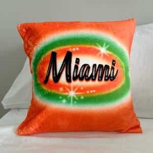 May include: Orange and green throw pillow with the word "Miami" in a black, stylized font. The pillow has a fuzzy texture.